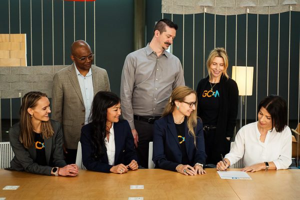 A group of people look at a woman who is signing a contract at a table.