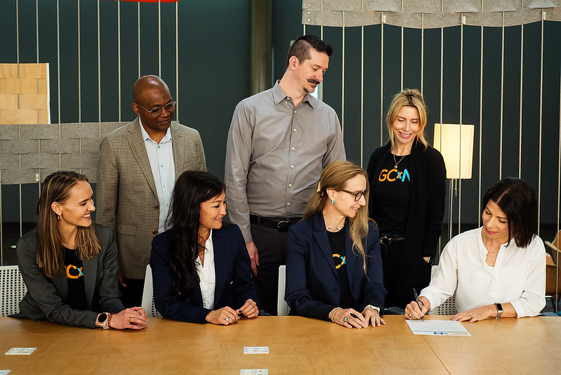 A group of people look at a woman who is signing a contract at a table.