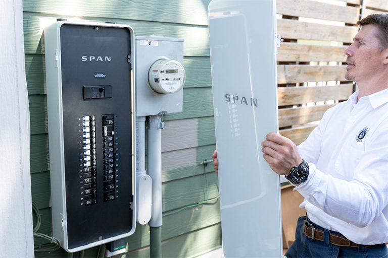 SPAN A man holds the cover of a power panel next to the panel hanging on the outside of a home.