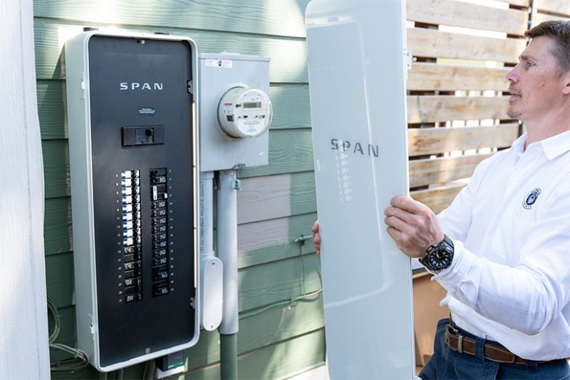 A man holds the cover of a power panel next to the panel hanging on the outside of a home.