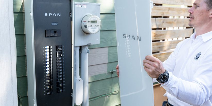 A man holds the cover of a power panel next to the panel hanging on the outside of a home.