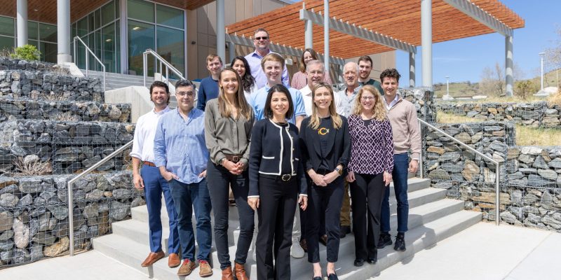 Fifteen people stand on steps outside a building and smile for the camera.