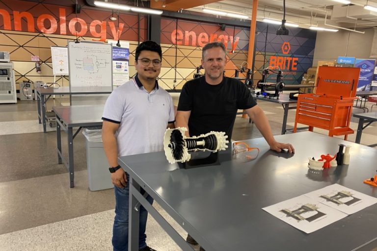 Two men stand in a lab setting with a piece of equipment in front of them