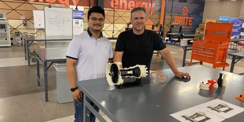 Two men stand in a lab setting with a piece of equipment in front of them