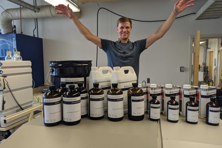 Evan with First Product to Ship 2023.03 Evan raises his arms, palms down, over a collection of containers with the name, “Zila Bioworks” written on them.