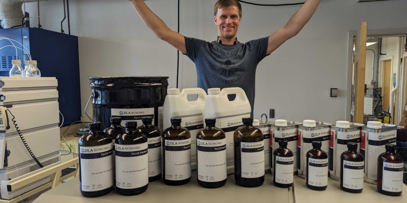 Evan raises his arms, palms down, over a collection of containers with the name, “Zila Bioworks” written on them.