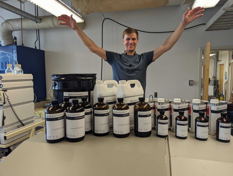 Evan raises his arms, palms down, over a collection of containers with the name, “Zila Bioworks” written on them.