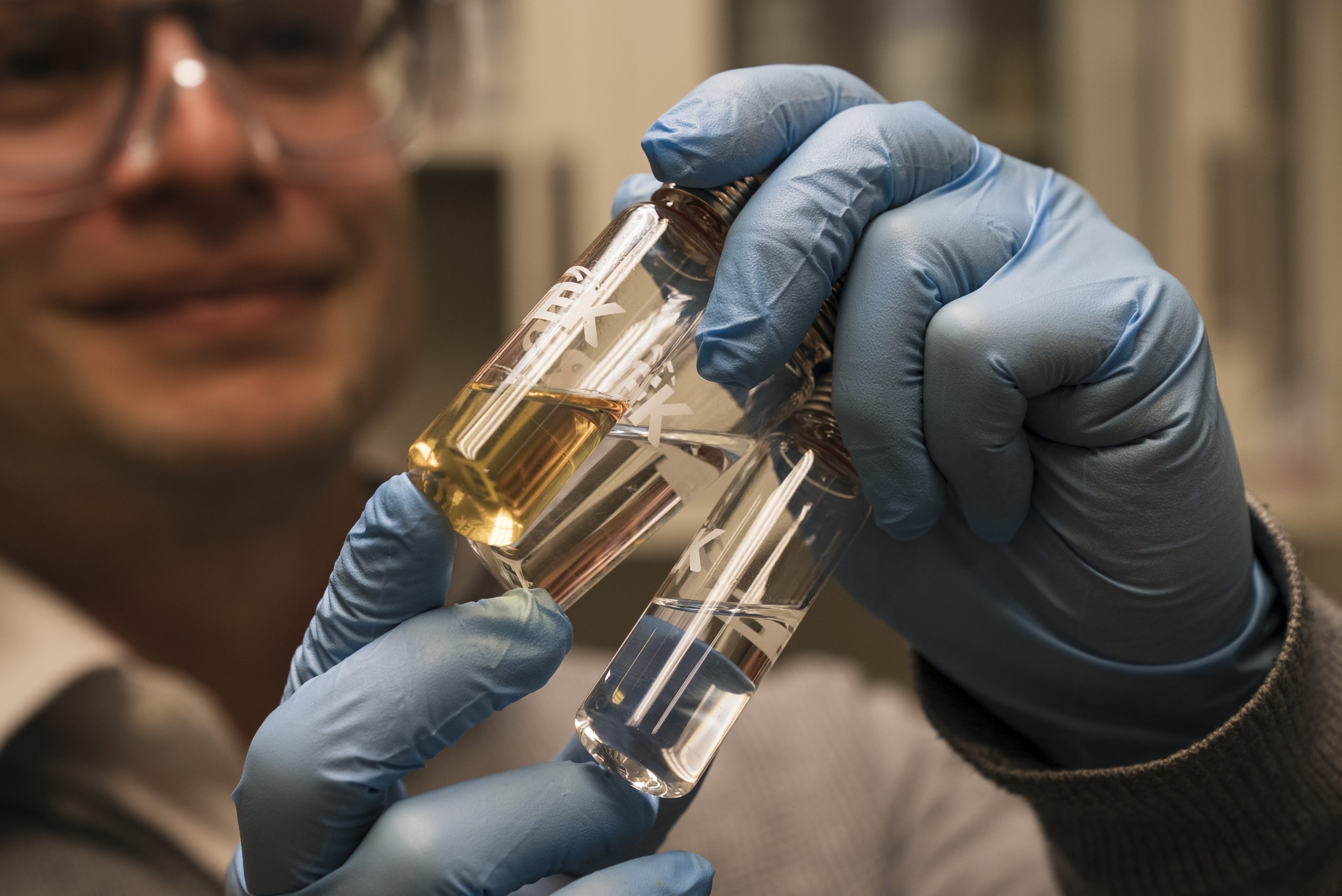 A person wearing blue gloves holds glass test tubes containing liquid samples.